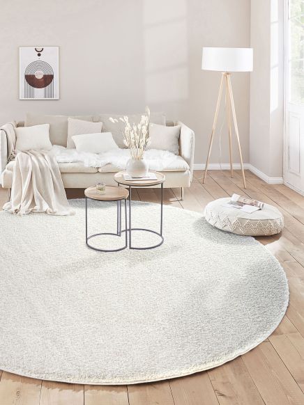 Beige Sofa mit Kissen und Decke vor runden Tischen auf weißem Teppich im hellen Wohnzimmer.