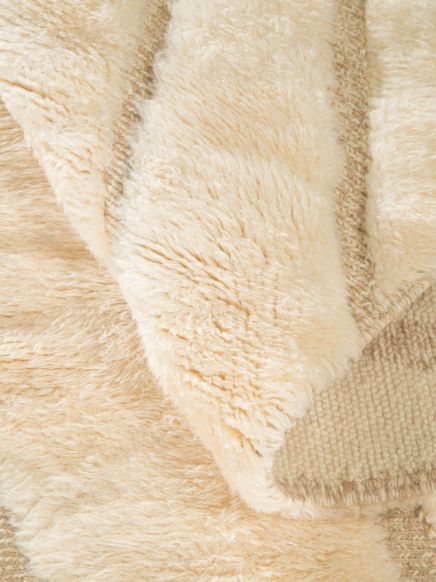 Wollteppich zeigt flauschige, cremefarbene Fasern, eine Ecke ist umgeklappt und offenbart die Rückseite.