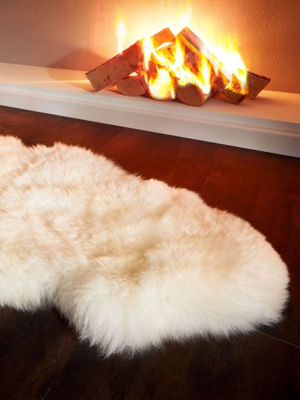 Fellteppich liegt auf dunklem Holzboden vor einem brennenden Kamin im Hintergrund