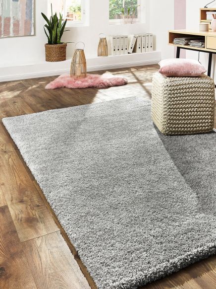 Grauer Teppich liegt auf Holzboden, daneben Pouf mit rosa Kissen in modernem Wohnraum.