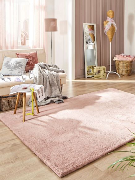 Kuscheliger rosa Teppich auf Holzfußboden neben Sofa mit Kissen und Decke rechts Spiegel und Schaufensterpuppe im Raum