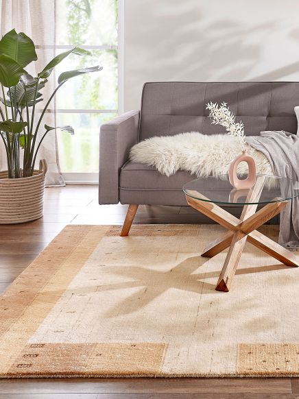 Grauer Sofa steht neben Glastisch mit Holzbasis im Wohnzimmer mit Teppich und Pflanze am Fenster