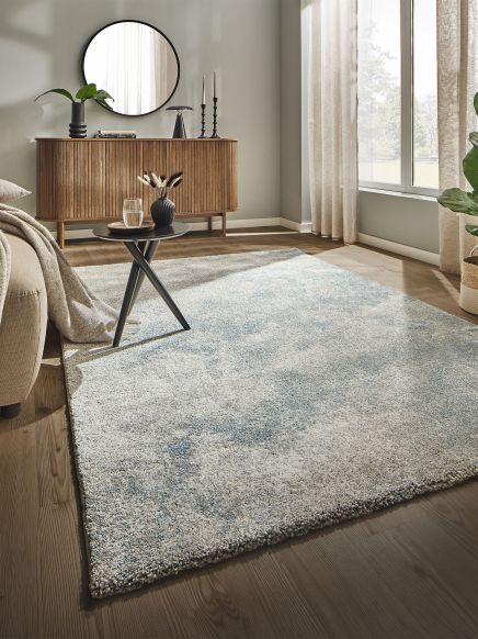 Heller Teppich liegt auf Holzboden, kleiner Tisch mit Vase und Glas, im Hintergrund Sideboard mit Spiegel.