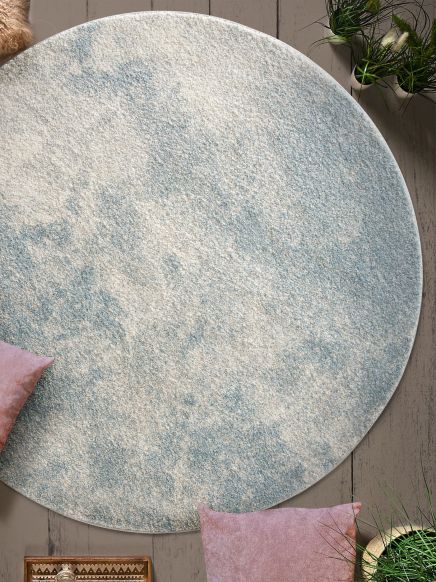 Runder Teppich mit weiß-blauem Muster liegt auf Holzboden, umgeben von rosa Kissen und Pflanzen.