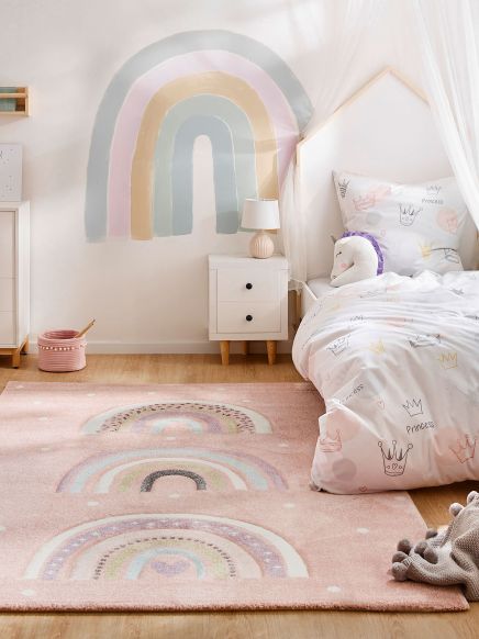 Kinderzimmer mit Regenbogen-Wandbild und Teppich ein Bett mit Einhorn-Kissen und Kronenmuster-Bettwäsche
