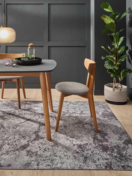 Stuhl steht neben Tisch mit Wasserkaraffe und Gläsern, Zimmer mit grauer Wand und Pflanze.