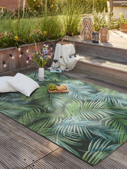 Teppich mit Blattmuster liegt auf Terrasse neben Kissen, Vase mit Blumen und Dekorationen