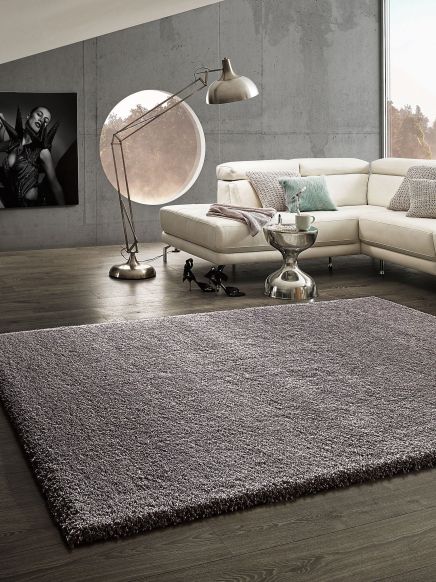 Großer grauer Teppich liegt auf dunklem Holzboden vor weißem Ecksofa mit Kissen im Wohnzimmer.