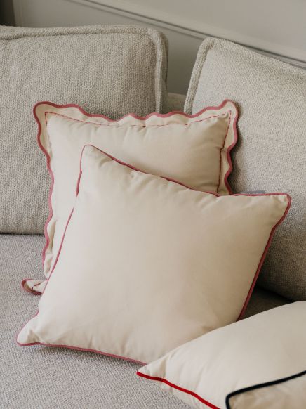 Zwei beige Kissen mit rotem Rand liegen auf einem cremefarbenen Sofa in einem Wohnzimmer.