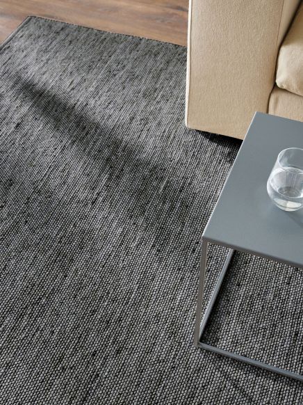 Grau melierter Teppich liegt unter grauem Beistelltisch mit Wasserglas neben beigem Sofa auf Holzboden.
