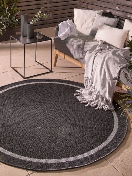 Runder Teppich liegt auf Terrasse neben Sofa mit Kissen und Decken ein Beistelltisch mit Vasen daneben